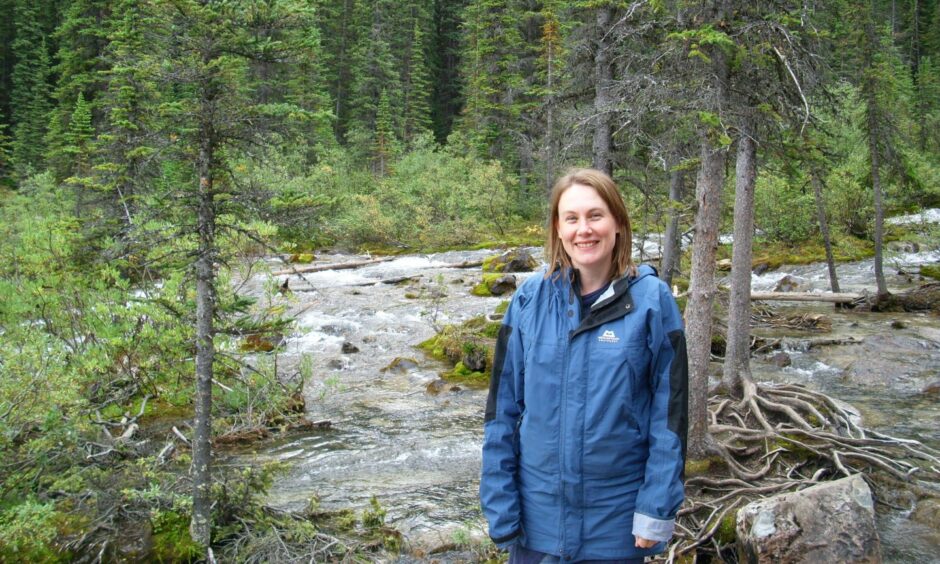 Sarah Field, smiling, wearing a blue waterproof jacket in a wooded area