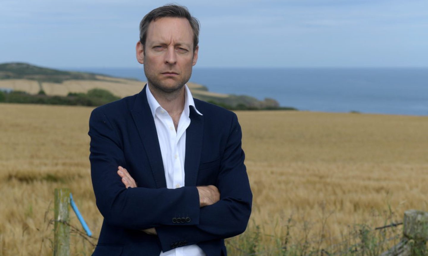 Liam Kerr in a suit with an open collar with his arms folded in a field. 