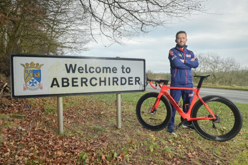 David Jarvis standing next to the "Welcome to Aberchirder" sign with his bicycle and Invictus Games attire on