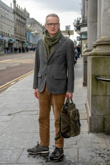 Filmmaker Robert Aitken on an Aberdeen street wearing brown trousers, a grey blazer and army green scarf.