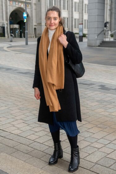 Aberdeen woman wearing a white turtleneck, blue midi skirt, black boots and coat, and a camel scarf.
