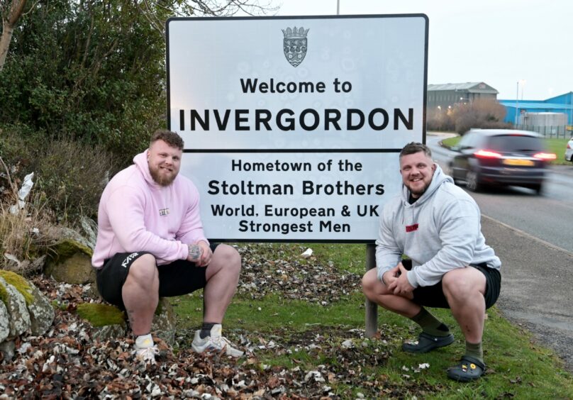 Tom and Luke Stoltman in front of road sign that reads, 'Welcome to Invergordon. Hometown of the Stoltman Brothers. World, European & UK Strongest Men.'