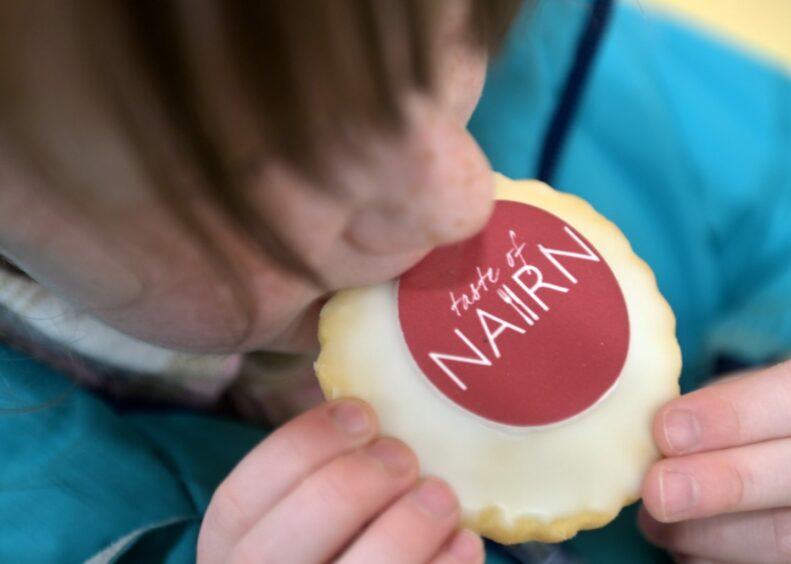 A girl in a turquoise jumper bites into a empire cake with a rice paper saying "Taste of Nairn". 