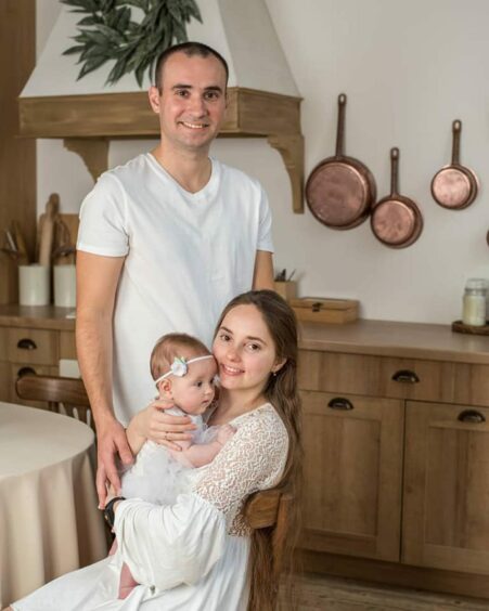 Ukrainian doctor Artur Yeroshenko is pictured standing behind his wife Yuliia, who is seated, holding their baby. They are all wearing white and shown in a rustic kitchen.