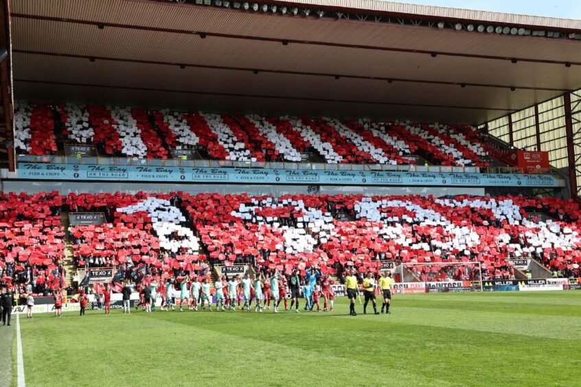 Aberdeen fans in the stands at Pittodrie Stadium with red and white displays that form the year "1983". Aberdeen and Hiberian players are walking on the pitch