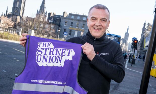 Bob Christie standing holding a purple hi-vis vest with the Our Union Street logo.