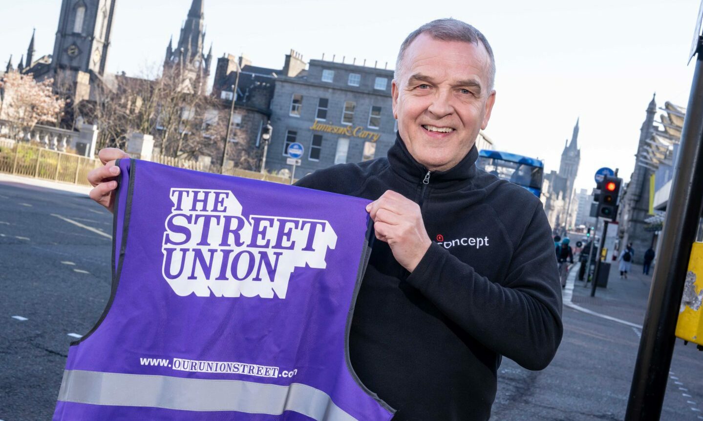 Bob Christie standing holding a purple hi-vis vest with the Our Union Street logo.