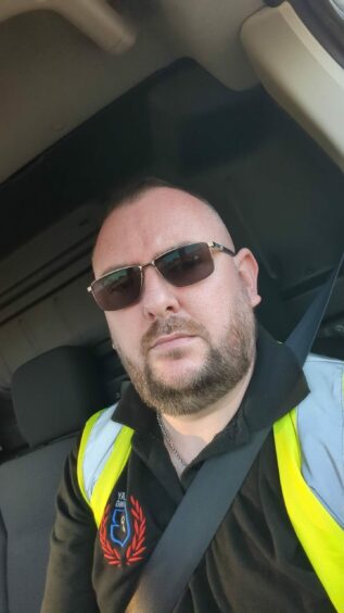Jon Barclay in the driving seat of a car with a high vis jacket on and wearing sunglasses. 
