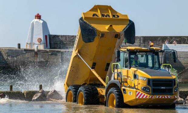 A yellow dumper truck viewed from the front with rocks being dropped into the water at the rear.
