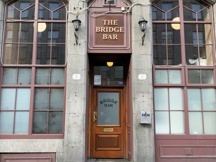 The high brown windows and traditional granite front of Aberdeen's Bridge Bar. Above the wooden door there is a sign saying 'the Bridge Bar.' Bridge Bar is also etched into the frosted window of the door. The bottom half of the window panes are steamed up. Spherical ceiling lights can be seen inside. 