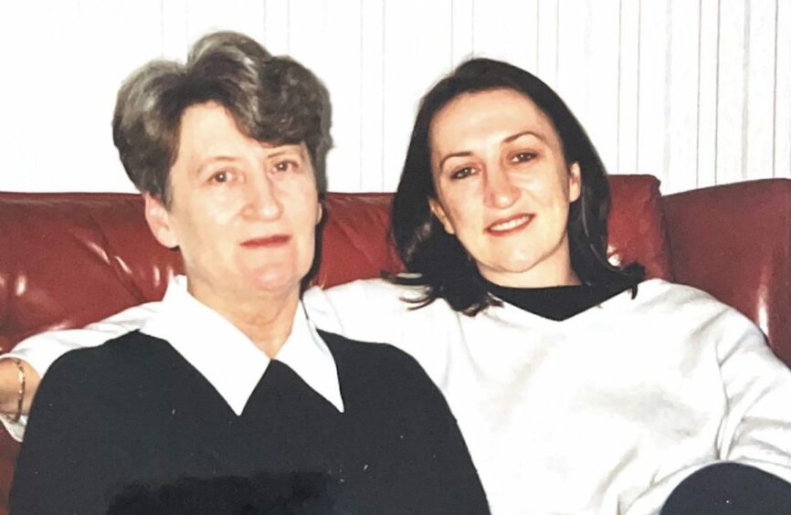 Sitting on a brown leather couch, mother Doris MacKenzie with short greying hair, a black jumper and white collar. Next to her is daughter Rhoda MacKenzie who has shoulder length black hair. She is smiling with her arm around her mum and is wearing a white V-neck jumper and black t-shirt.