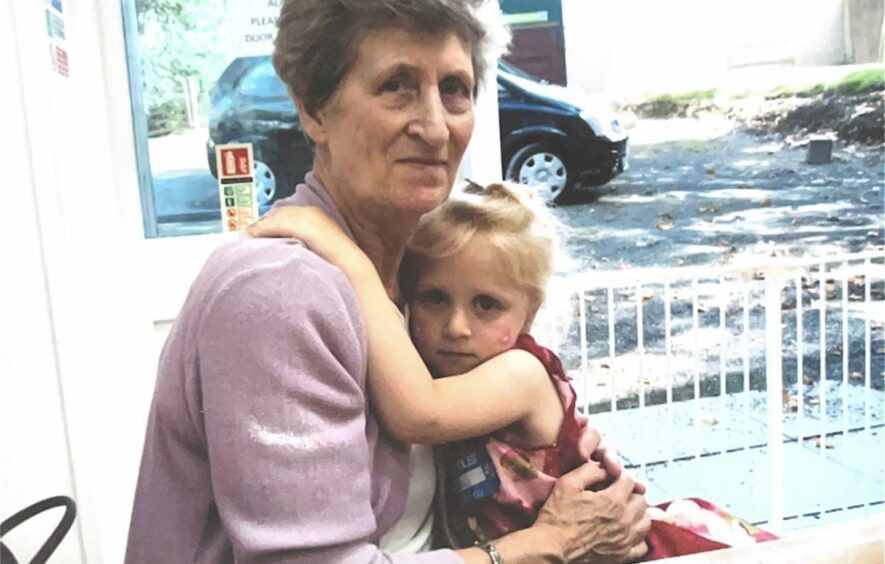 Grandmother Doris MacKenzie is wearing a lilac cardigan and cream blouse. Her hair is short and grey. On her lap, with arms around Doris' neck, is granddaughter Maia. She has a fair complexion, blonde hair and is wearing a pink and red sleeveless dress. 