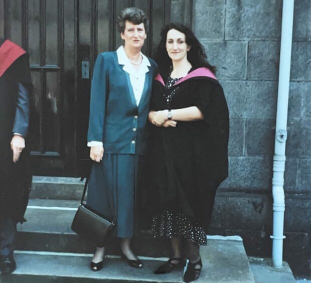 Wearing a formal blue long skirt and matching jacket with a white blouse is Doris MacKenzie. Tall and slim with dark hair she is standing in front of a granite wall and black wooden doors next to daughter Rhoda. Rhoda is wearing a patterned dress over which there is a black graduation gown and pink hood. Image taken on Rhoda's graduation day.