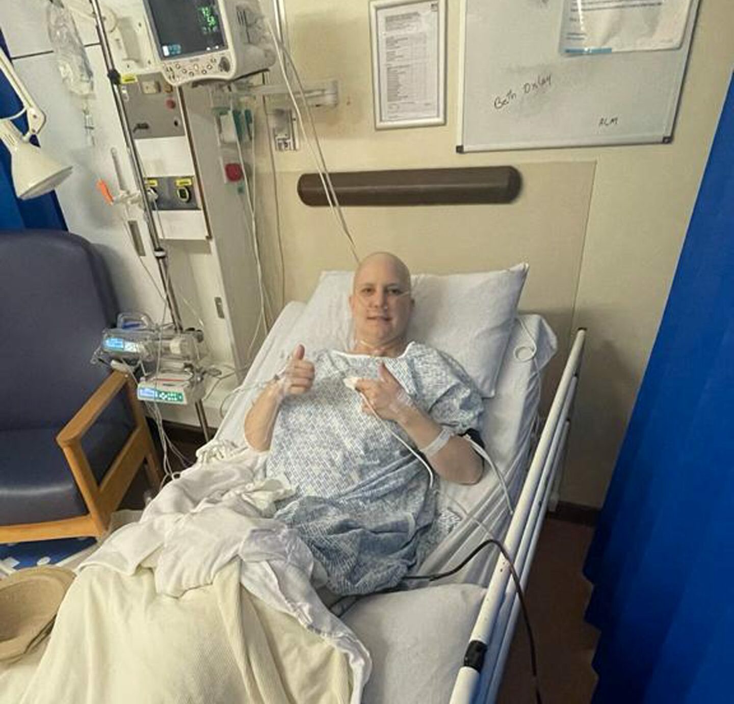 Face-on view of Beth Oxley in a hospital bed connected to various machines with her thumbs up. 