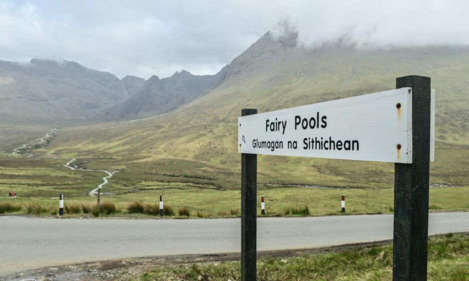 The sign for the car park at the Fairy Pools. A white sign in the foreground reads Fairy Pools, with the name written in Gaelic as well. 