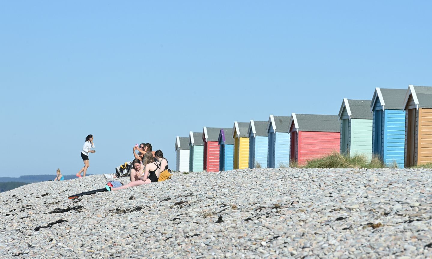 A small group of people sit on the beach in front of some colourful beach huts with not a cloud in the sky. 