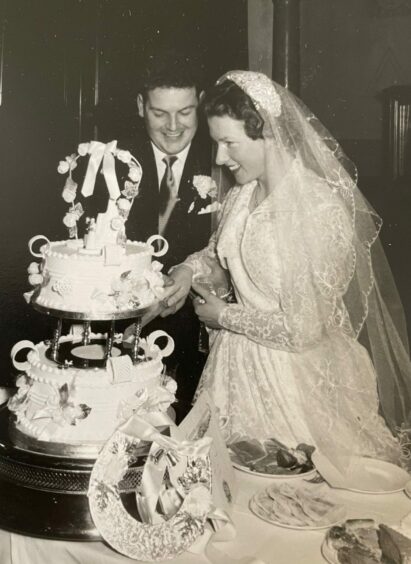 John Ramage and his bride Jean cutting their wedding cake. It's a black and white photo. She is wearing an intricately detailed lace dress and veil, John is in a dark with a carnation in his top pocket. Both are smiling.