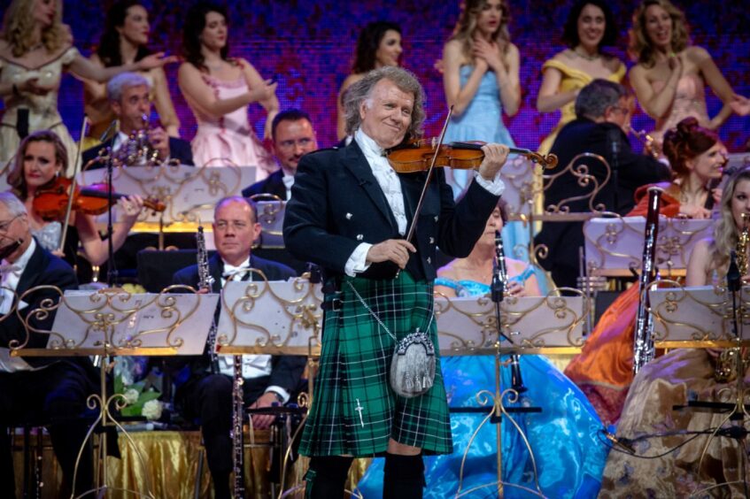 Andre Rieu playing the violin with a smile on his face wearing a green kilt, fastened with a sword-styled kilt pin and wearing a Prince Charlie jacket and waistcoat and a grey sporran. 