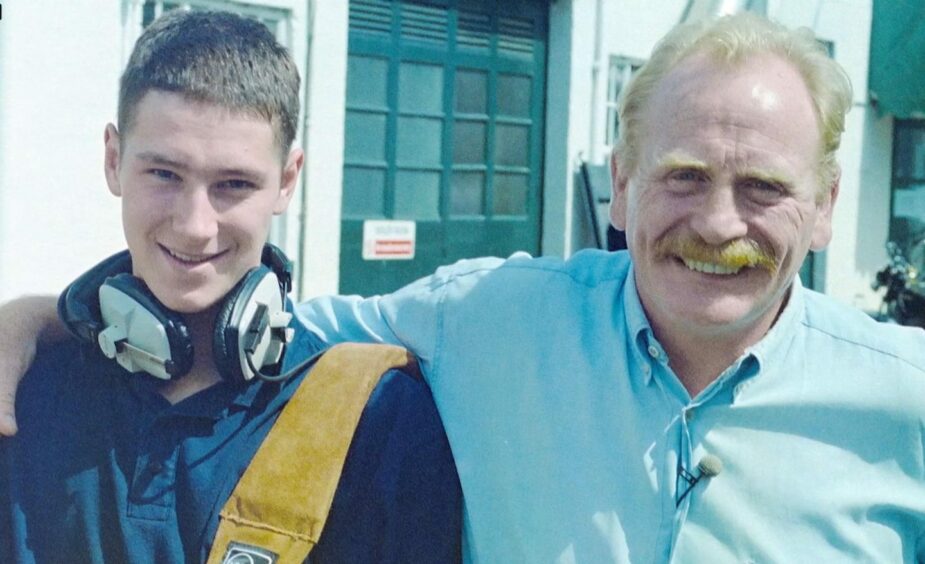 A young man - Ranald Wood - is pictured wearing a blue polo shirt with headphones around his neck, smiling. He is next to Braveheart actor James Cosmo who is also smiling, wearing a pale blue shirt, hugging Ranald. 