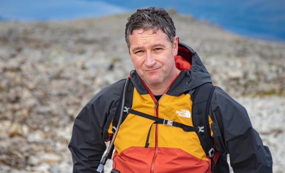 Pictured at the summit of Ben Nevis, Ranald Wood is shown wearing a black, orange and yellow The North Face waterproof jacket. His hair is greying, and his rucksack straps are visible. 