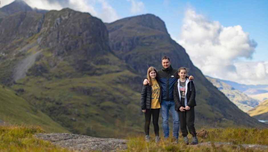 With the mountains of Glencoe in the background Ranald is shown in the foreground wearing a waterproof jacket and jeans with his two nieces, one at either side of him. He has his arms around their shoulders. Everyone is smiling. 