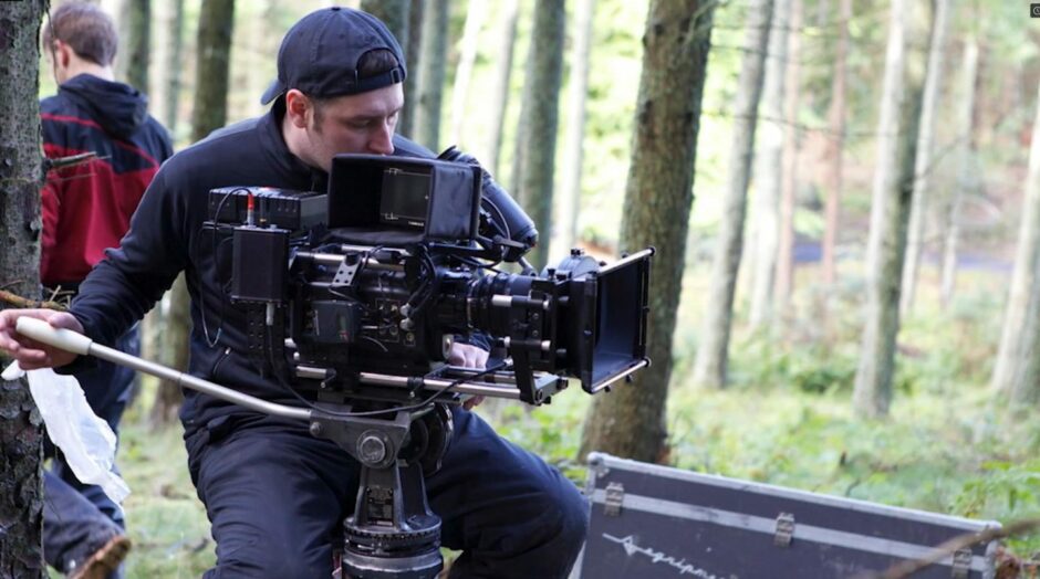 Wearing a back to front baseball cap, sitting on a stool and looking into a movie camera, Ranald Wood, is pictured filming in a woodland.