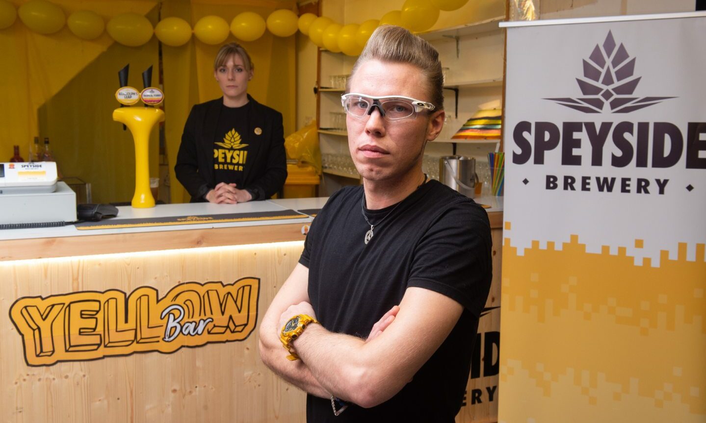 Seb Jones with arms folded standing in front of a bar with Yellow Bar branding, and a sign with Speyside Brewery branding. 