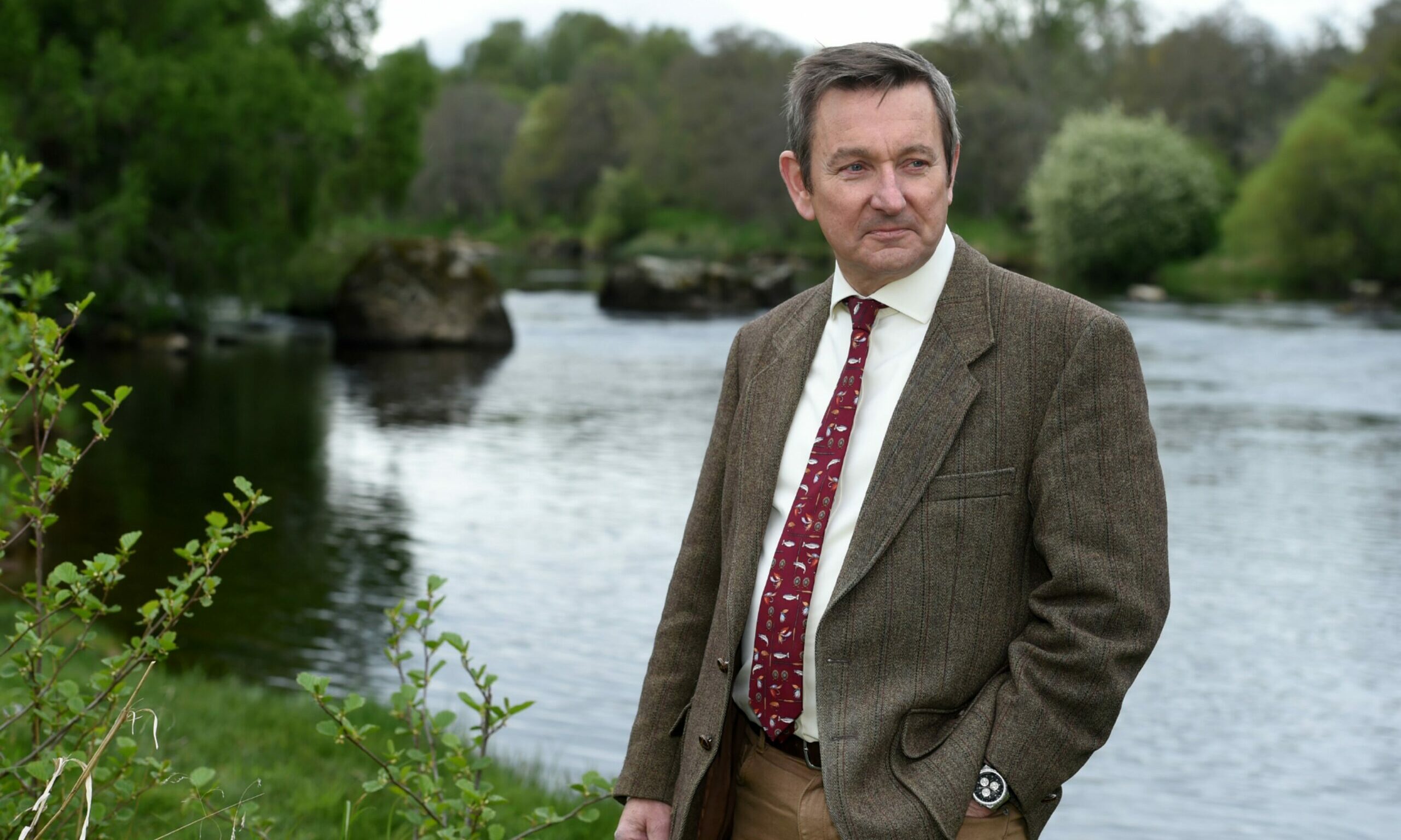 Roger Knight wearing tweed on bank of River Spey looking to camera right with one hand in pocket. 