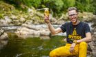Seb Jones holds a glass of beer while kneeling on banks of a river.