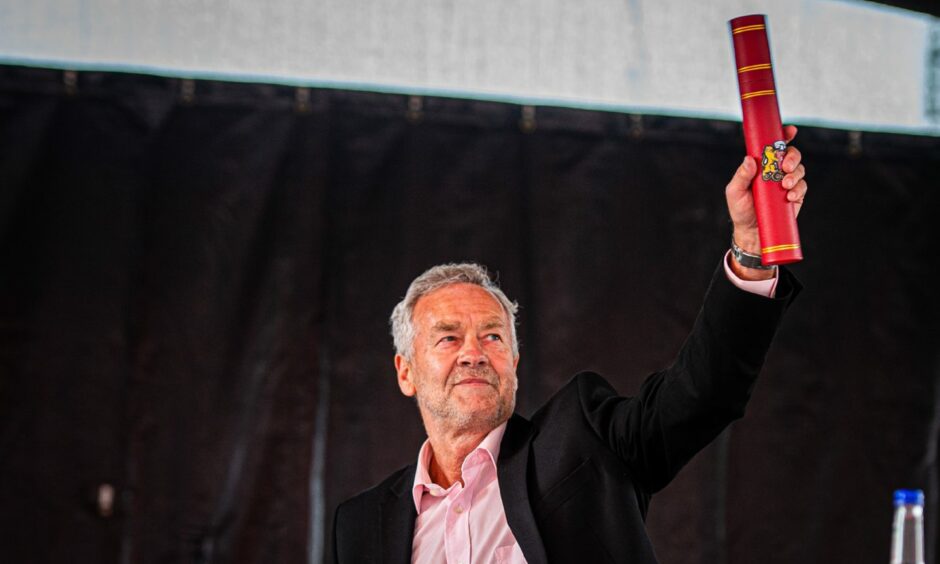 Dougie Bell holds high his award. Mr Bell is wearing a pink shirt and dark jacket and his hair if grey, he is smiling and the award is in a red box. 