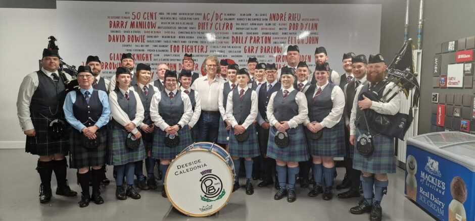 Members of the Deeside Caledonia Pipe Band standing with Andre Rieu holding their bagpipes, with a large drum that reads "Deeside Caledonia".
