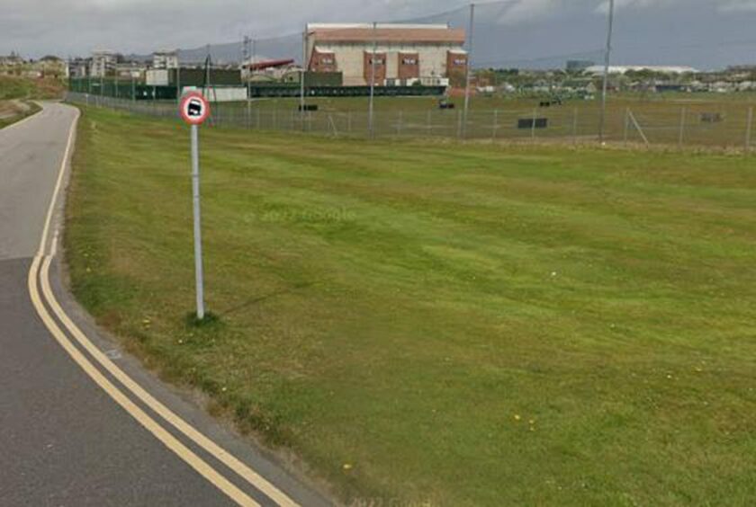 A screenshot of the grass area next to Accommodation Road with a sign in the foreground showing a car half parked on the road and Pittodrie Stadium in the background. 