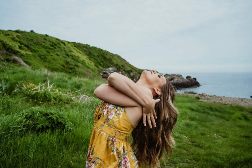 Emma leaning back with her arms around herself on a hillside with the sea behind her