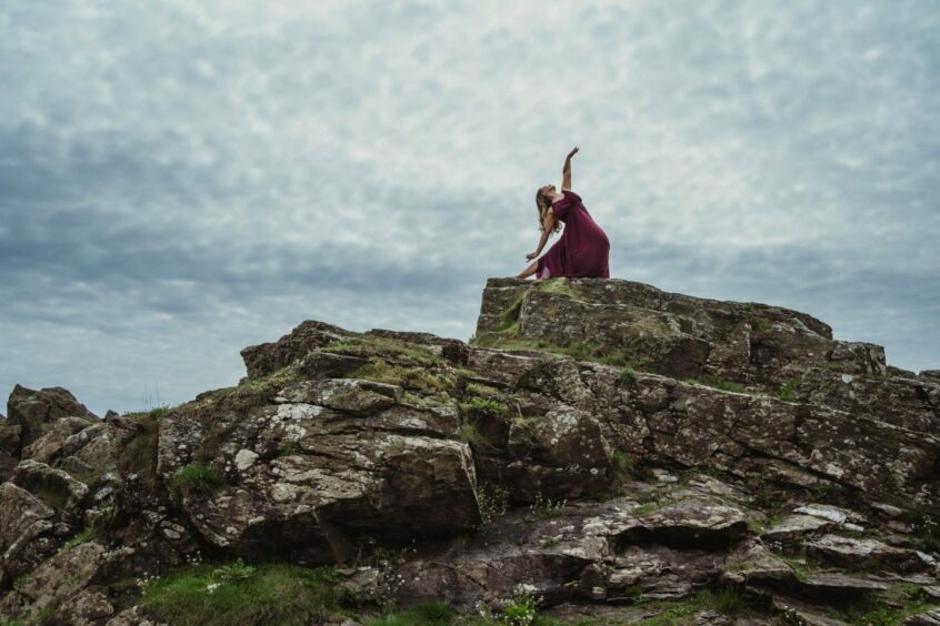 Emma at the top of a rocky hill, lunging with one of her arms reaching to the sky