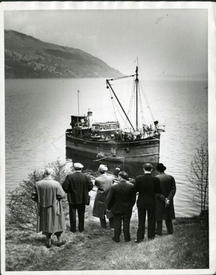 A black and white picture showing the backs of six well-dressed men and women as they look towards a puffer boat on Loch Ness
