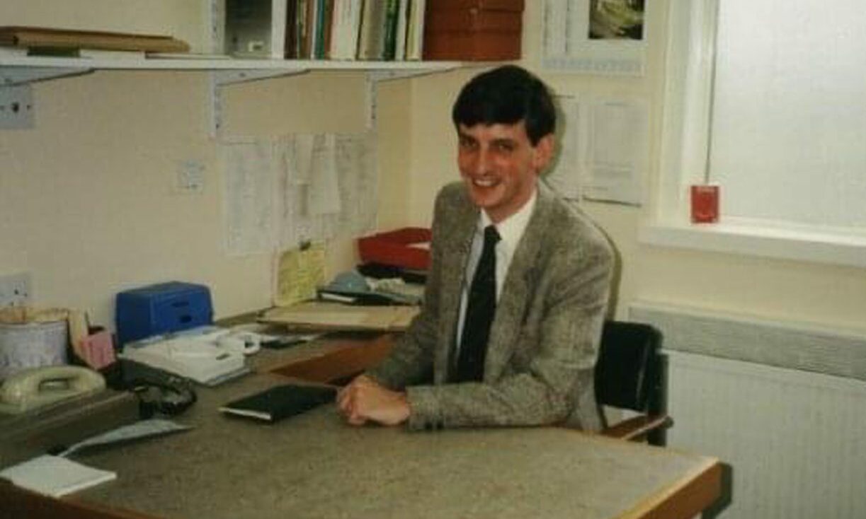 A young Iain Small sitting behind a desk in his GP office smiling at the camera. 
