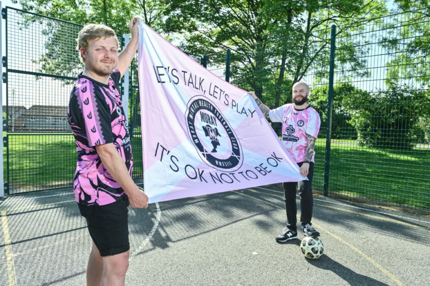 Co-founders Phil Barton and Matty Slinger holding a flag that reads "let's talk, let's play. It's okay not to be okay" with the Moray Mental Health Football Club logo on it