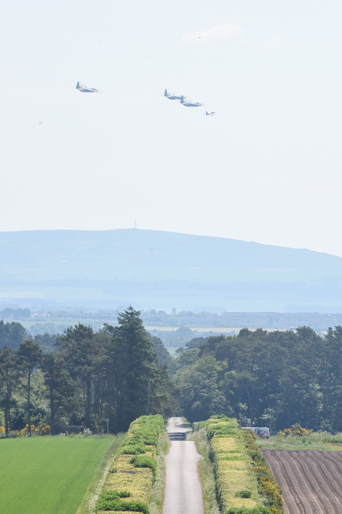 Wide shot of three Hercules planes banking to the right with a Typhoon jet over the countryside. 