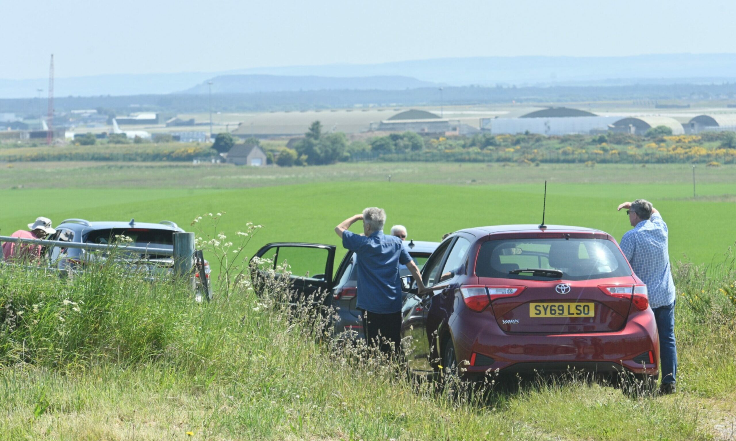Cars lined up in a field with drivers looking to the sky with RAF Lossiemouth in the background. 