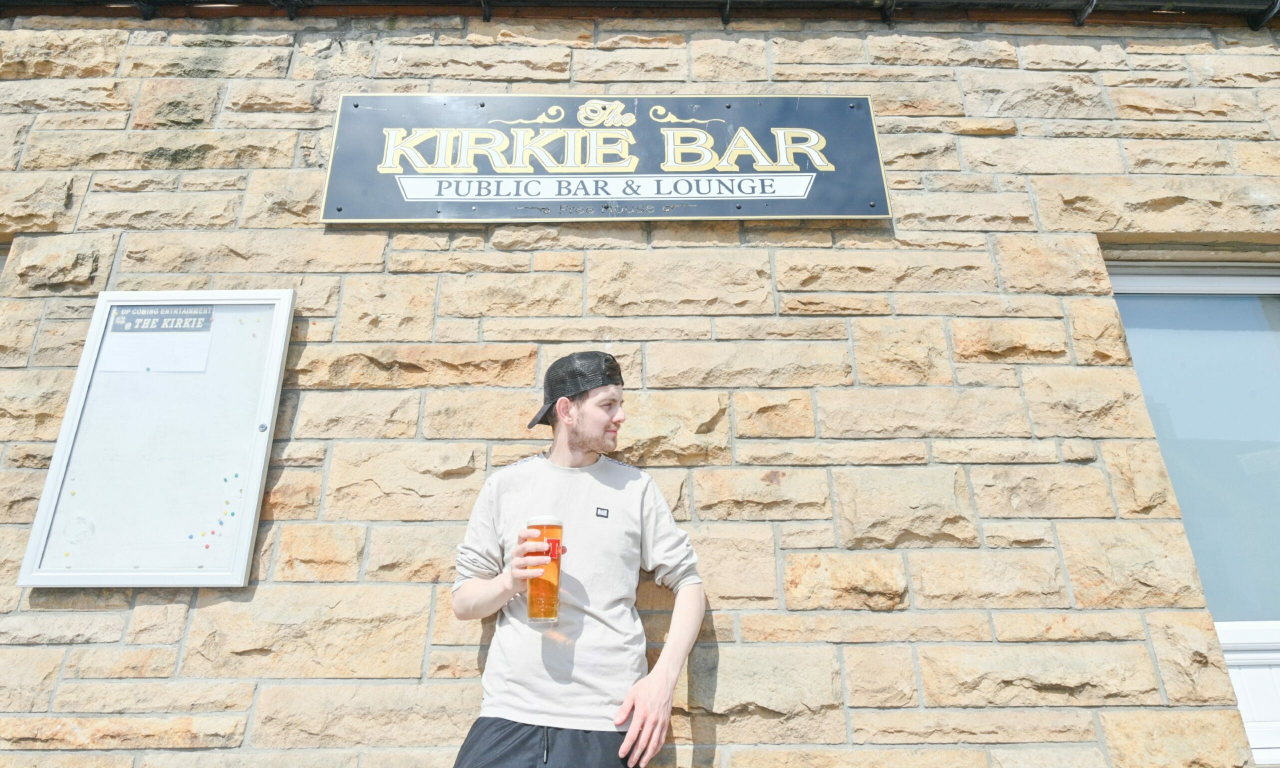 Danny Grant standing outside Kirkie with back to wall looking to camera's right holding pint of Tennent's. 