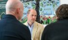 Prince William talking to people viewed between the heads of two people from behind.