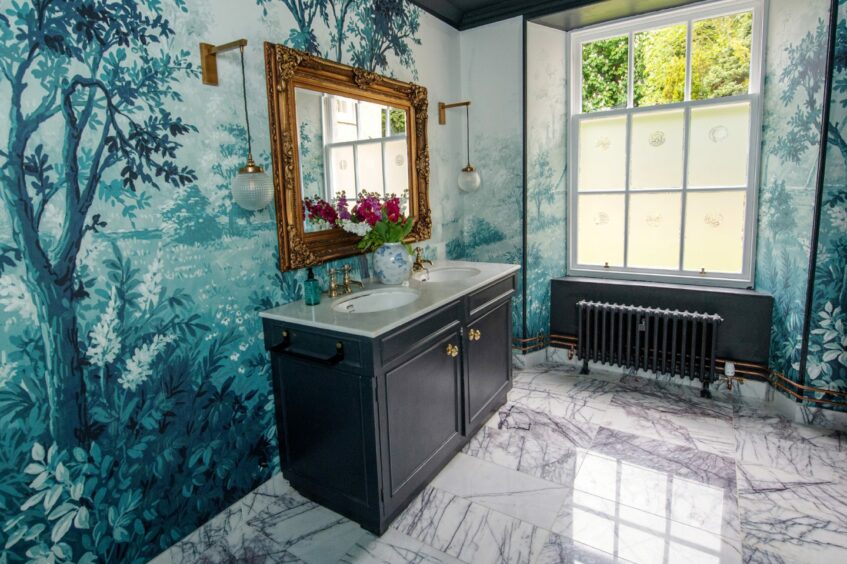 A double sink with a vase of flowers on it and a gold framed mirror hung above and two hanging lights on either side. The room has marble floor tiles, forest print wallpaper and a large frosted window