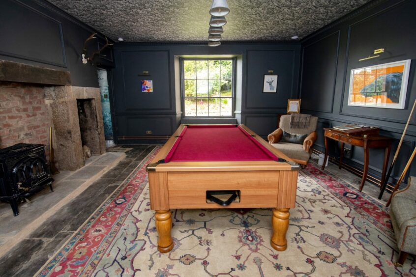 A rustic looking room with stone flooring, a patterned rug, pool table, fireplace and armchairs.