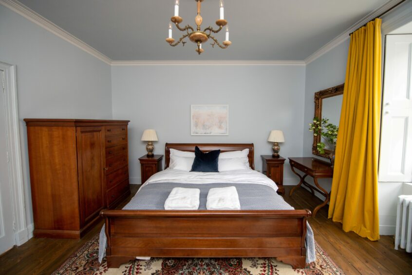 One of the bedrooms of the house, decorated with dark wood tones, pale blue walls and bedding and yellow curtains