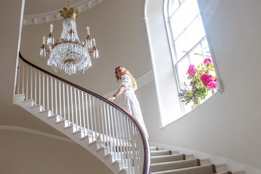 Christina Horspool walking up the curved stairway, an electric chandelier above her and a vase of flowers on the windowsill behind her