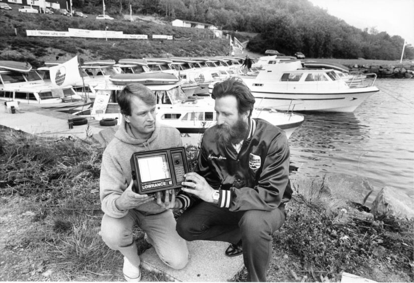 Adrian Shine and David Stiensland holding electronic equipment on the shores of Loch Ness with about a dozen small boats moored behind them.