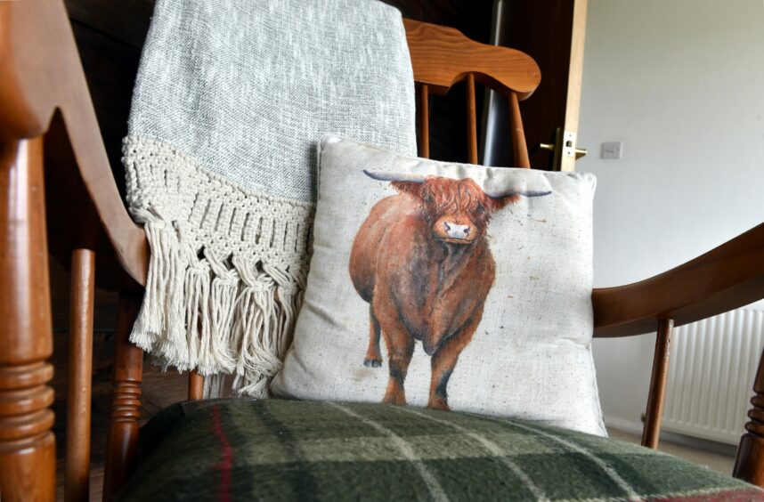 A wooden chair with tartan upholstery, a cushion with a highland cow printed on it and a throw hanging over the back of it.