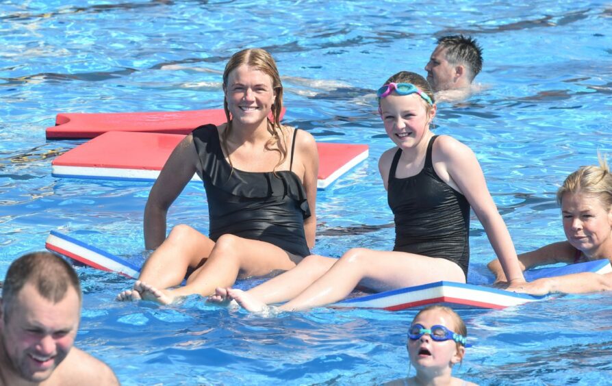 A woman and girl sit on paddle board as people swim round them.