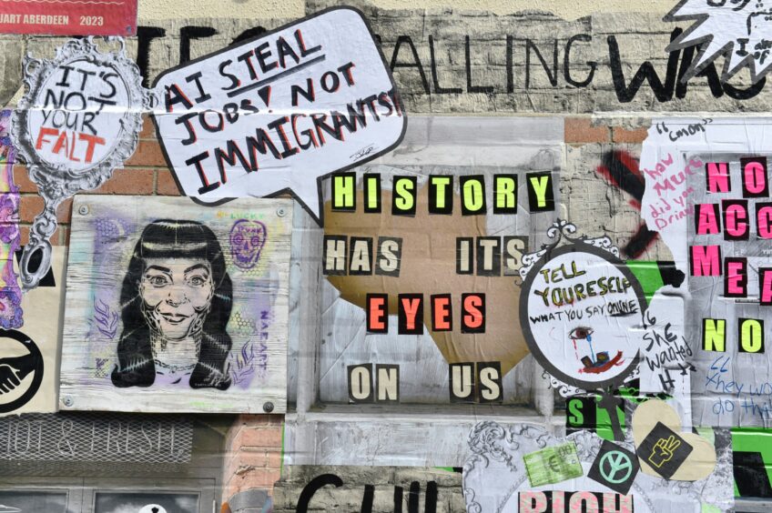 A brick wall covered in painted posters and slogans, with the main one reading "History has its eyes on us" in yellow, beige and orange print.