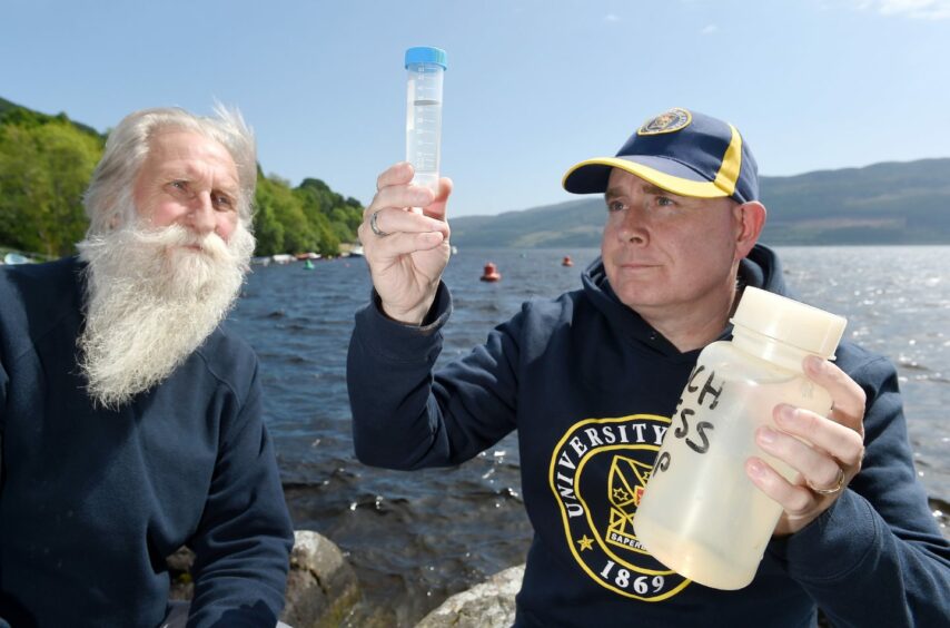 Two men, one with a long white beard, looking at a glass sample container on the shores of Loch Ness